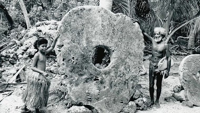 Two people on the island of Yap stand beside a large circular rai stone, a traditional form of money whose ownership could be transferred even though the stone itself was too heavy to move.