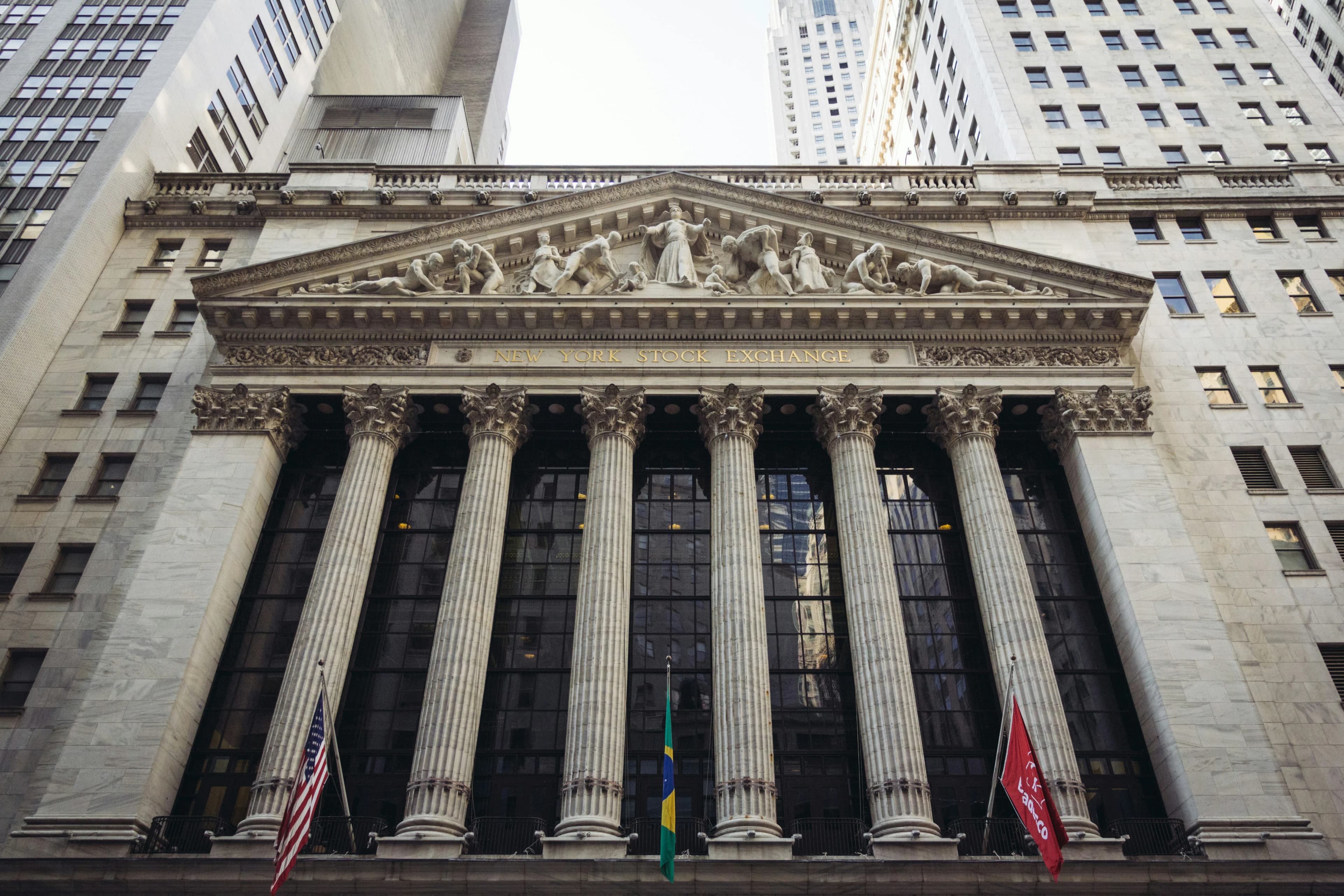 The neoclassical facade of the New York Stock Exchange building at 11 Wall Street, fronted by six tall Corinthian columns and a sculpted pediment, with a large American flag draped across the front of the building.